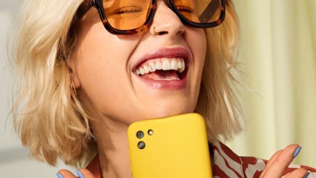 Lächelnde Frau mit großer Sonnenbrille hält ein gelbes Smartphone in der Hand.