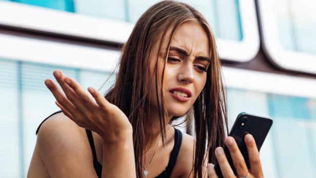 Junge Frau schaut mit verwirrtem Gesichtsausdruck auf ihr Smartphone und hebt fragend die Hand.
