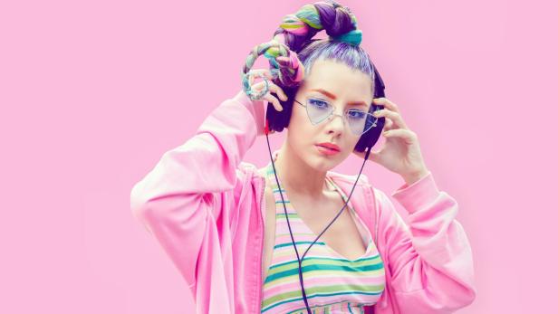 Junge Frau mit buntem Zopf, Kopfhörern und modischer Brille trägt gestreiftes Top und pinke Jacke vor rosa Hintergrund.
