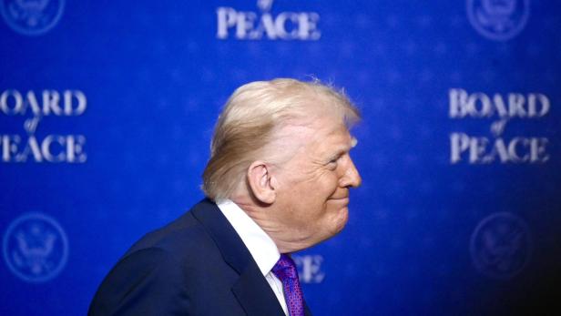 Ein Mann im Anzug mit blonden Haaren steht seitlich vor blauem Hintergrund mit der Aufschrift „Board of Peace“.