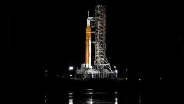 Eine große Rakete steht nachts beleuchtet auf einer Startrampe, umgeben von Scheinwerfern und Gerüsten.