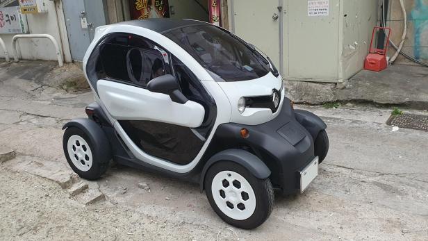 Ein weiß-schwarzer Renault Twizy steht auf einer leicht beschädigten Straße vor einem Gebäude.