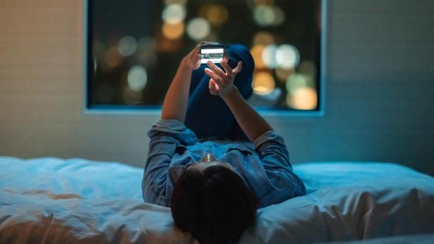 Person liegt abends auf dem Bett und benutzt ein Smartphone, im Hintergrund leuchten unscharfe Stadtlichter.