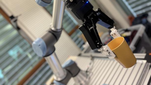 Roboterarm mit Greifhand hält einen gelben Becher in einem Laborraum.