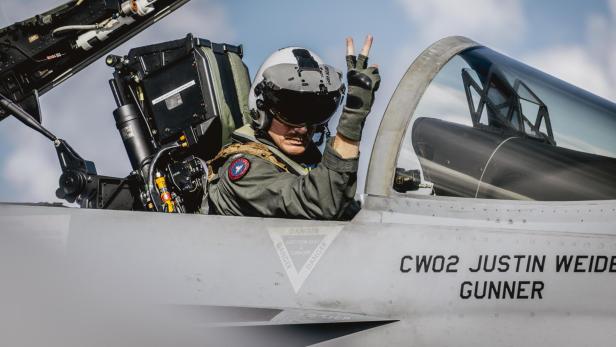 Pilot mit Helm und Handschuhen zeigt Victory-Zeichen im Cockpit eines Kampfjets bei offenem Dach.
