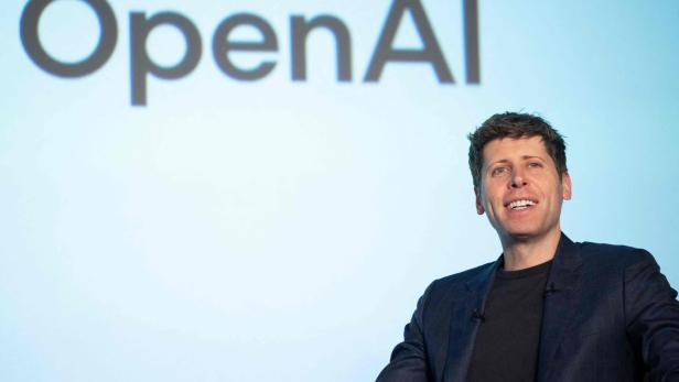 Ein Mann lächelt vor dem "OpenAI"-Schriftzug auf hellblauem Hintergrund.