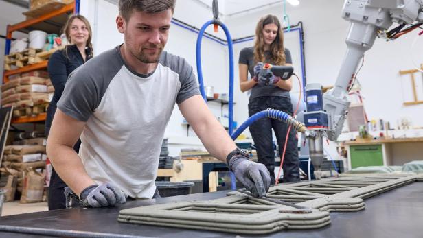 Drei Personen arbeiten mit einem 3D-Betondrucker an einem Fachwerkträger in einer Werkstatt.