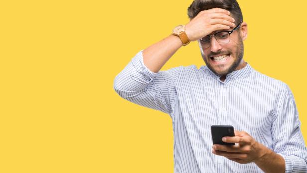 Ein Mann mit Brille und Hemd blickt entsetzt auf sein Handy vor gelbem Hintergrund.