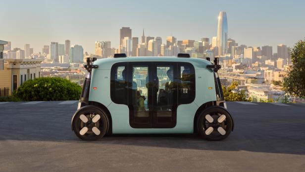 Autonomes Zoox-Robotaxi vor Skyline einer US-Großstadt bei Tageslicht.