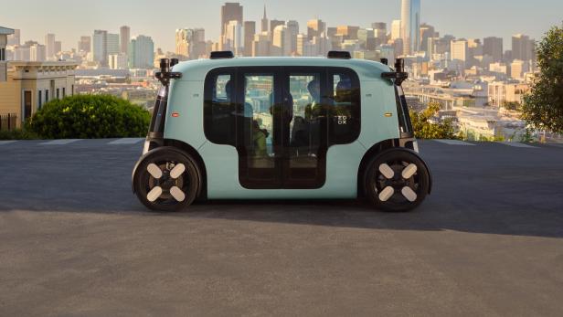 Autonomes Zoox-Robotaxi vor Skyline einer US-Großstadt bei Tageslicht.