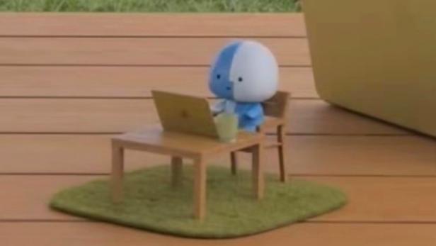 Apple-Maskottchen Lil Finder Guy sitzt auf einem Mini-Schreibtisch auf einem richtigen Tisch