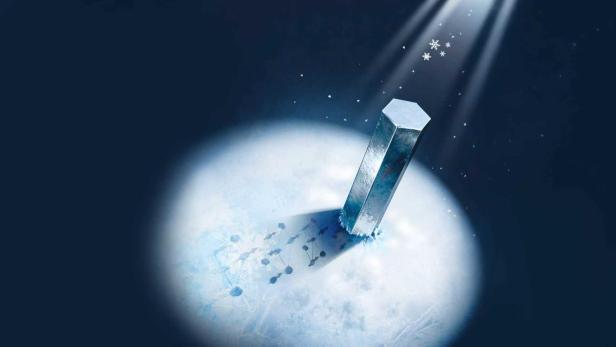 Ein silberner Metallblock steht auf einer vereisten Fläche, umgeben von Schneeflocken und angestrahlt von Licht, symbolisiert die ultrakalte Legierung EuCo2Al9.