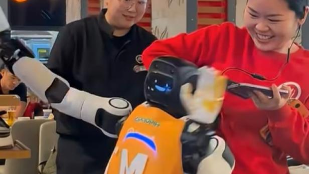 ein humanoider Roboter mit den Händen in der Luft wird von einer Frau im roten Pulli am kragen gepackt