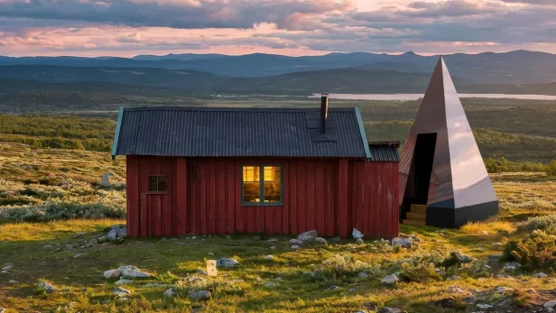 Rotes Holzhaus und eine moderne pyramidenförmige Struktur in hügeliger Landschaft bei Sonnenuntergang.