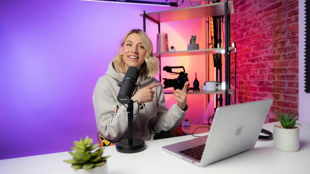 Eine Creatorin sitzt vor Mikrofon und Laptop in einem modernen Studio und spricht, im Hintergrund Kamera-Equipment und bunte Beleuchtung.