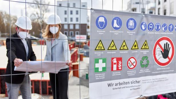 Zwei Personen mit Schutzhelmen und Masken betrachten Baupläne hinter einem Bauzaun mit Warnhinweisen.