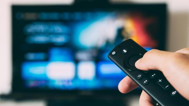 Hand hält Fernbedienung vor unscharfem Fernsehbildschirm mit Streaming-Menü.