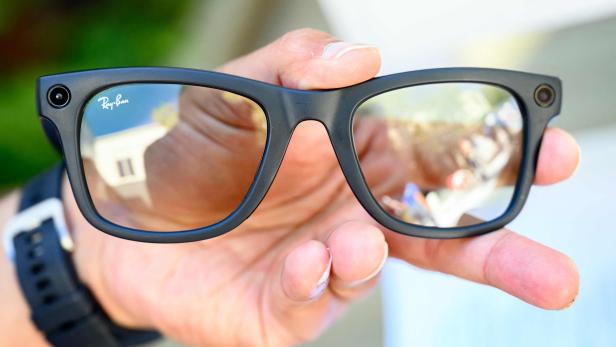 Eine Hand hält eine schwarze Ray-Ban-Brille mit integrierten Kameras an den Ecken des Rahmens.