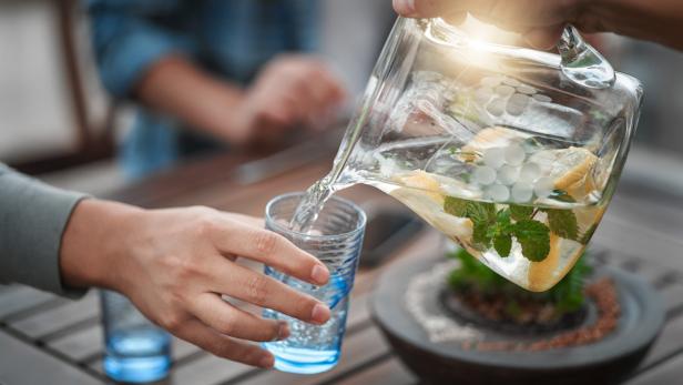 Wasser mit Zitronenscheiben, Minze und Eiswürfeln wird aus einer Glaskaraffe in ein Trinkglas gegossen, um auf die Bedeutung von Wasserqualität im Alltag hinzuweisen.