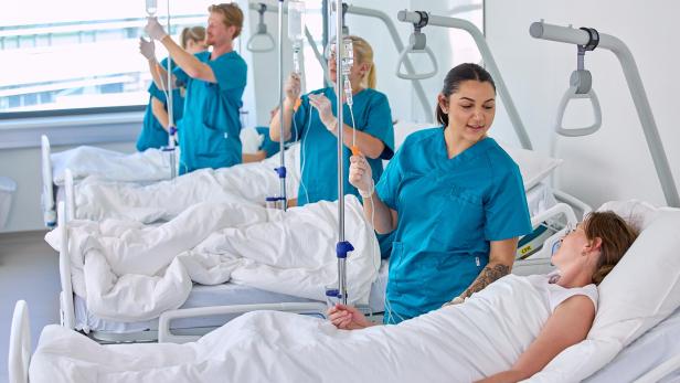 Pflegekräfte betreuen mehrere Patientinnen und Patienten in Krankenhausbetten und legen Infusionen an.