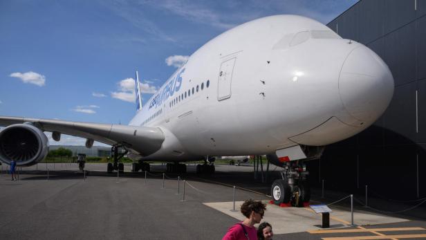 Ein Airbus A380 steht auf einem Flugplatz, während zwei Personen im Vordergrund vorbeigehen.