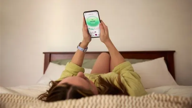 Frau liegt am Bett mit Garmin-Uhr und geöffneter Natural Cycles App am Smartphone.