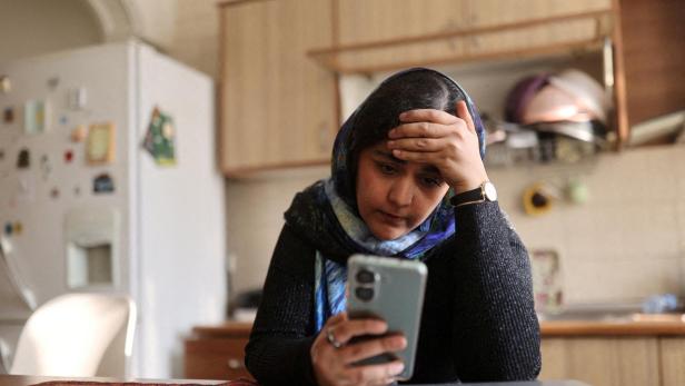 Eine Frau mit Kopftuch sitzt nachdenklich am Tisch, hält ein Smartphone in der Hand und stützt den Kopf ab.