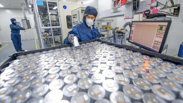 Eine Person in Schutzkleidung überprüft zahlreiche zylindrische Batteriezellen in einer modernen Fabrikumgebung.