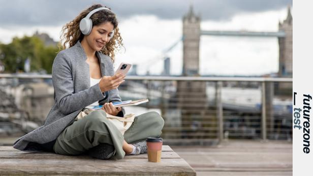 Frau mit Kopfhörern nutzt ein Smartphone im Freien, im Hintergrund die Tower Bridge.