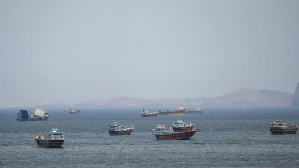 Mehrere Schiffe liegen auf ruhigem Meer, im Hintergrund sind bergige Küsten zu sehen.