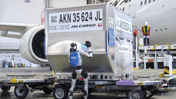 Ein humanoider Roboter mit blauem Rucksack bewegt einen großen Frachtcontainer neben einem Flugzeug in einer Lagerhalle.