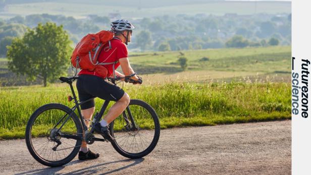 Ein Mann mit Helm und rotem Rucksack fährt bei sonnigem Wetter auf einem Fahrrad durch eine grüne Landschaft.