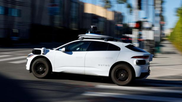 Ein weißes autonomes Auto mit Sensoren auf dem Dach fährt durch eine städtische Straße bei Tageslicht.