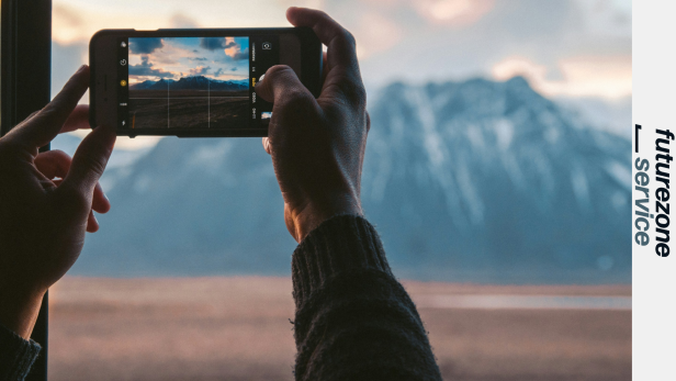 Person fotografiert eine Berglandschaft mit dem iPhone bei Sonnenuntergang durch ein Fenster.