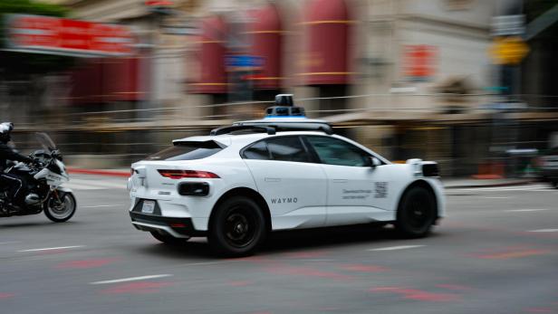 Ein weißes autonomes Auto mit Sensoren auf dem Dach fährt neben einem Polizeimotorrad durch eine Stadtstraße.