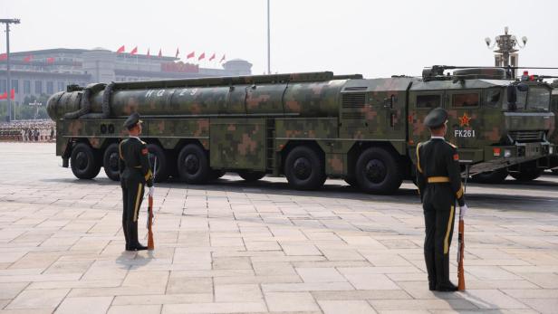 Großer Militärlastwagen mit Tarnmuster und Rakete, flankiert von zwei Soldaten mit Gewehren auf einem Platz.