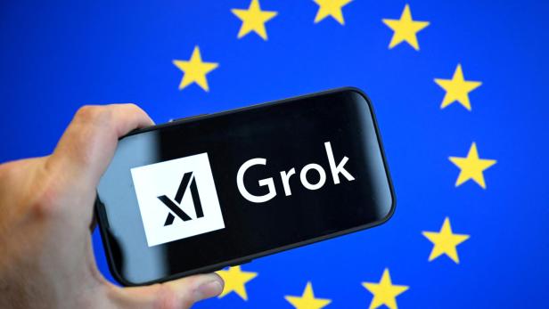 Eine Hand hält ein Smartphone mit dem Grok-Logo vor einem Hintergrund mit gelben Sternen auf blauem Grund.