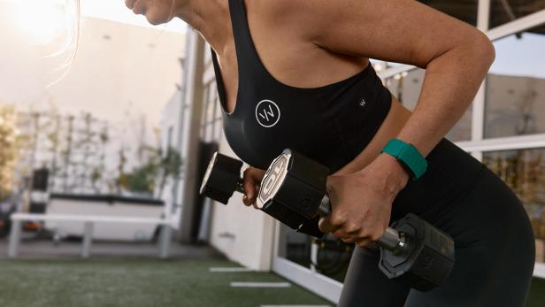 Eine Frau im Sportoutfit hebt Hanteln, an ihrem linken Handgelenk ein grünes Whoop-Band