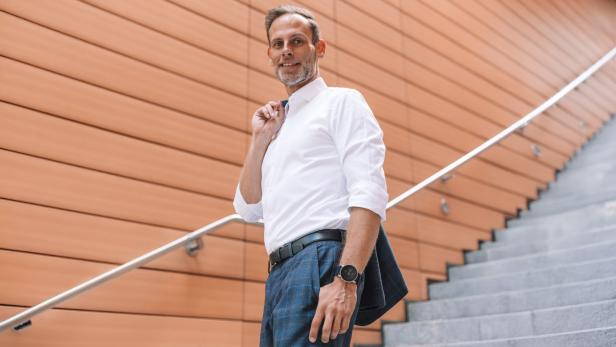 Ein Mann in Businesskleidung steht auf einer Treppe vor einer modernen Wandverkleidung und lächelt.