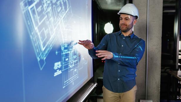 Fachmann im Baustellenhelm zeigt auf digitale Architekturpläne in einem modernen Büro.