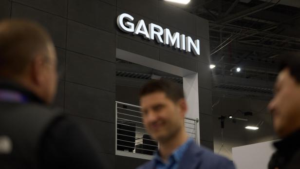 Drei unscharfe Personen stehen vor einem Messestand mit großem leuchtendem Garmin-Schriftzug und moderner Architektur.