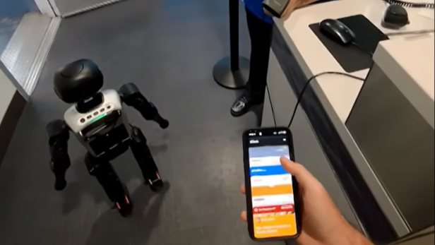 Ein humanoider Roboter steht an einem Flughafenschalter neben einer Person mit Smartphone.