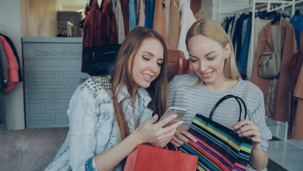 Zwei Frauen sitzen nebeneinander, eine hält ein Smartphone, die andere eine gestreifte Einkaufstasche