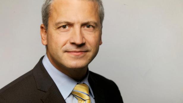 Mann mit grauen Haaren im Anzug und gelber Krawatte vor neutralem Hintergrund.
