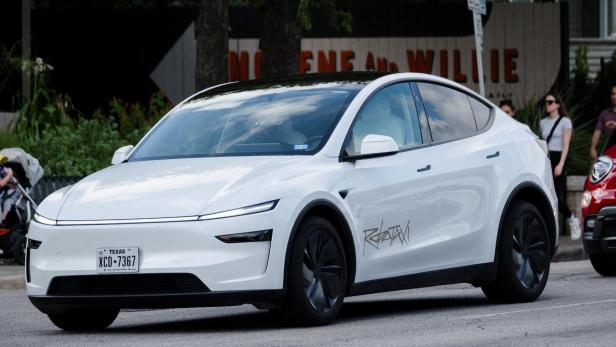 Ein weißes Elektroauto mit schwarzen Felgen und getönten Scheiben fährt auf einer Straße, im Hintergrund sind Fußgänger zu sehen.