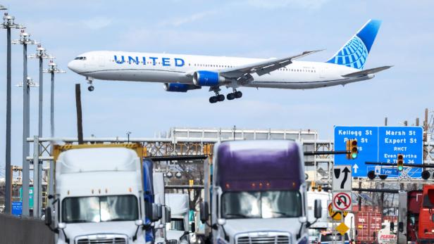 Ein Flugzeug fliegt niedrig über einer belebten Straße mit mehreren Lastwagen und Verkehrsschildern.