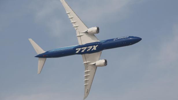 Ein großes blau-weißes Passagierflugzeug mit der Aufschrift „777X“ fliegt schräg am Himmel.