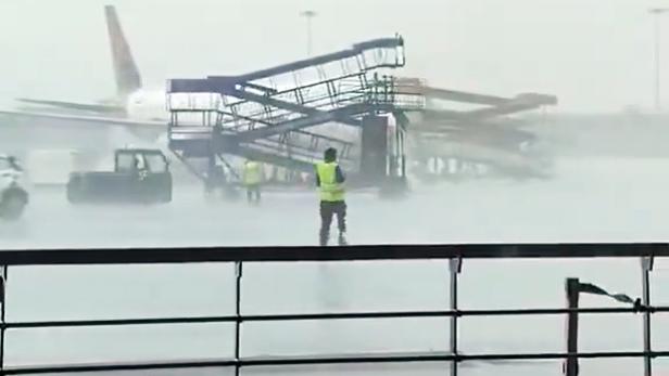 Die Gangway rollt über den Flughafen, das Wetter ist regnerisch und windig. Im Hintergrund steht ein Flugzeug