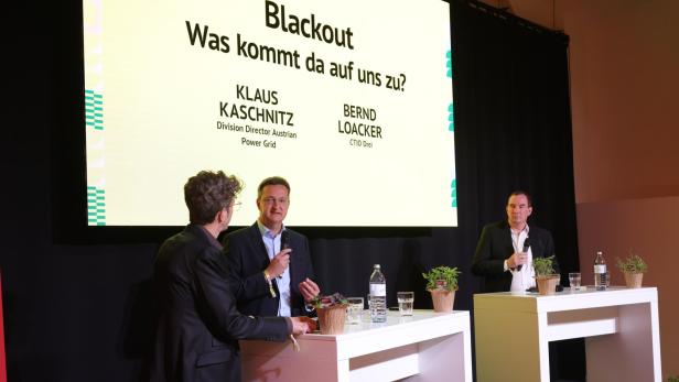 Drei Männer im Anzug diskutieren auf einer Bühne mit Mikrofonen vor einer großen Präsentationsfolie zum Thema Blackout.