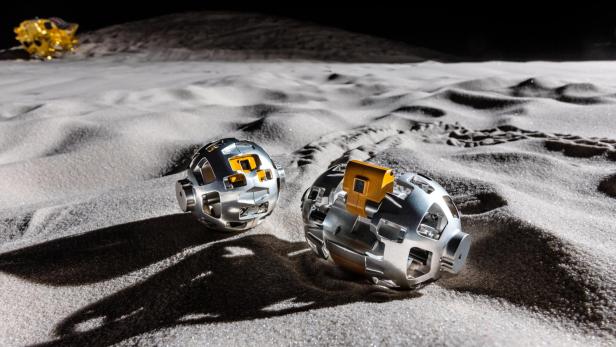 Zwei silberne, kugelförmige Roboter mit gelben Elementen liegen auf sandigem Untergrund in einer simulierten Mondlandschaft.